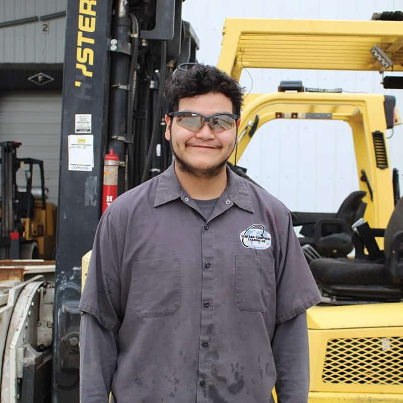 A man wearing safety glasses and a gray work shirt stands and smiles in front of a yellow forklift outside an industrial building.