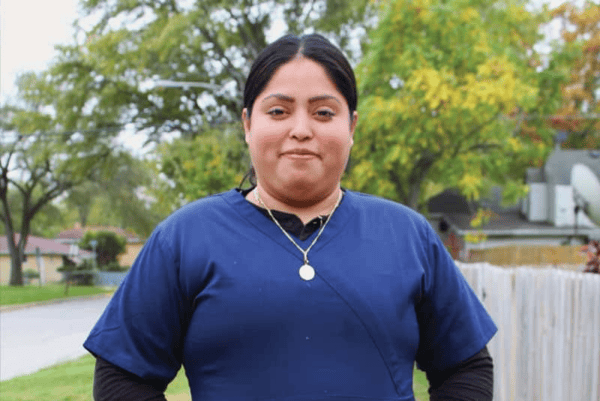 A woman wearing blue medical scrubs and a black undershirt stands outdoors on a sidewalk, with her hands in her pockets. Trees and houses are visible in the background. She is looking at the camera and smiling slightly.