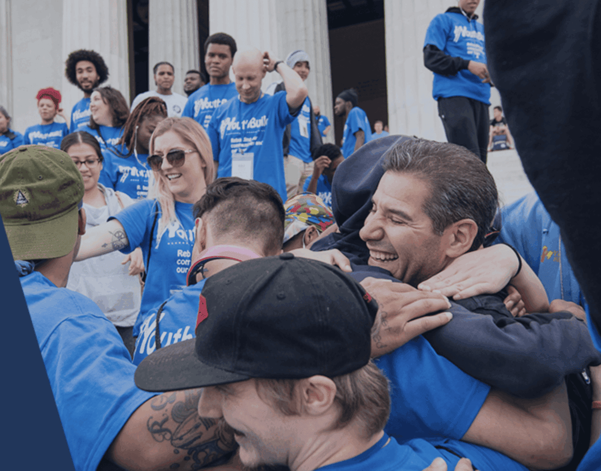 A diverse group of people wearing blue shirts gather outdoors on steps, smiling and hugging each other in a celebratory, joyful atmosphere.