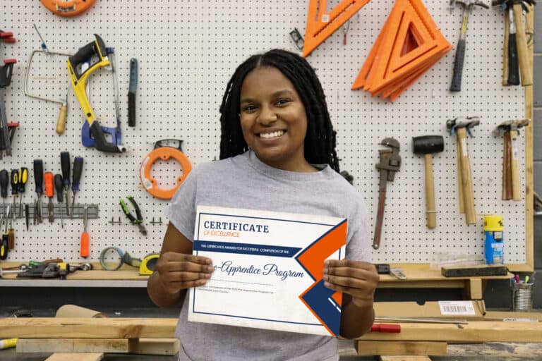 A smiling woman stands in a workshop, holding a certificate. Various tools, such as hammers and wrenches, hang on a pegboard behind her, along with wood and measuring triangles.